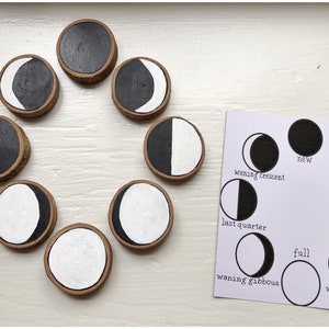 May include: A set of eight wooden moon phases painted in black and white. The phases are arranged in a circle on a white surface. A card with the names of the moon phases is shown to the right of the wooden moon phases.