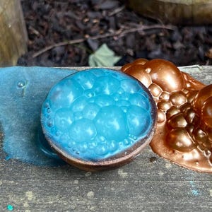 May include: Three bath bombs with textured surfaces. One is teal, one is blue, and the other two are bronze. The bath bombs are on a wooden surface. The teal bath bomb has a small amount of teal powder around it.