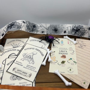 K&ouml;nnte beinhalten: Ein Halloween-Bastelset mit verschiedenen Materialien. Das Set enth&auml;lt bedruckte Karten mit gruseligen Illustrationen, k&uuml;nstliche Knochen, eine schwarze Spinne und dekoratives Band mit Spinnweben und Totenk&ouml;pfen. Das Set ist zum Basteln gedacht.