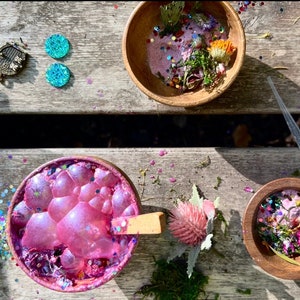 May include: A wooden bowl filled with pink slime, glitter, and dried flowers. A wooden spoon is in the bowl. A second wooden bowl is filled with dried flowers and glitter. A clear plastic dropper is on the table.