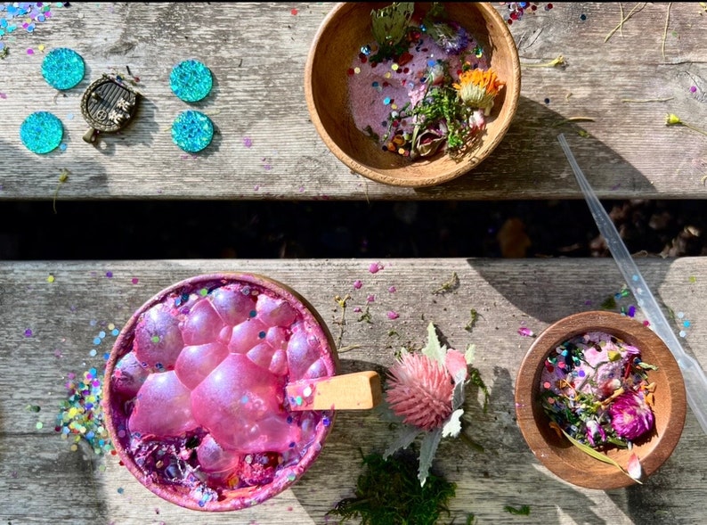 May include: A wooden bowl filled with pink slime, glitter, and dried flowers. A wooden spoon is in the bowl. A second wooden bowl is filled with dried flowers and glitter. A clear plastic dropper is on the table.