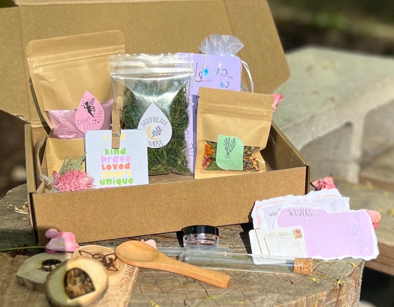 May include: A brown cardboard box filled with various items, including a bag of pink powder labeled "Pink Fairy Dust", a bag of green moss labeled "Moonbeam Moss", a bag of dried flowers labeled "Garden Herbs", a wooden spoon, a small glass jar with glitter, a wooden heart with a fairy design, and two purple cards with the words "Love & Wishes" written on them.