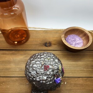 K&ouml;nnte beinhalten: Eine strukturierte, silberfarbene Kerze mit getrockneten Blumen und Glitzer liegt auf einer Holzoberfl&auml;che. Eine bernsteinfarbene Glasflasche mit Korken und eine kleine Holzschale mit violetten Granulaten sind ebenfalls vorhanden.