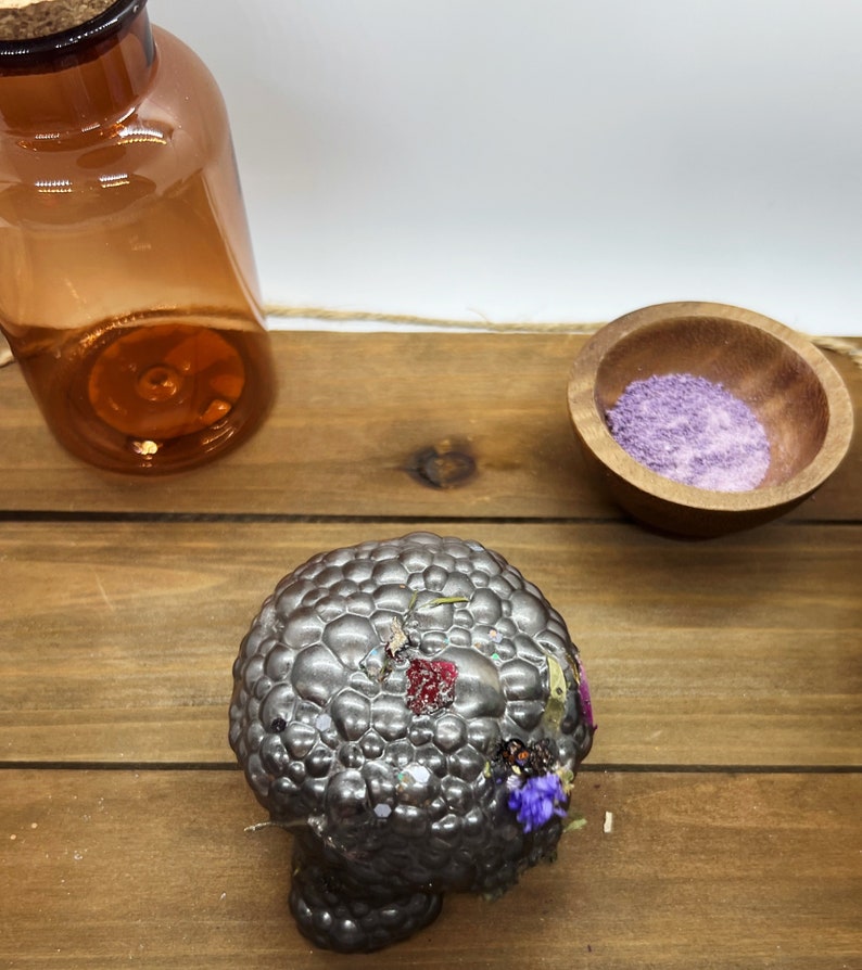 K&ouml;nnte beinhalten: Eine strukturierte, silberfarbene Kerze mit getrockneten Blumen und Glitzer liegt auf einer Holzoberfl&auml;che. Eine bernsteinfarbene Glasflasche mit Korken und eine kleine Holzschale mit violetten Granulaten sind ebenfalls vorhanden.