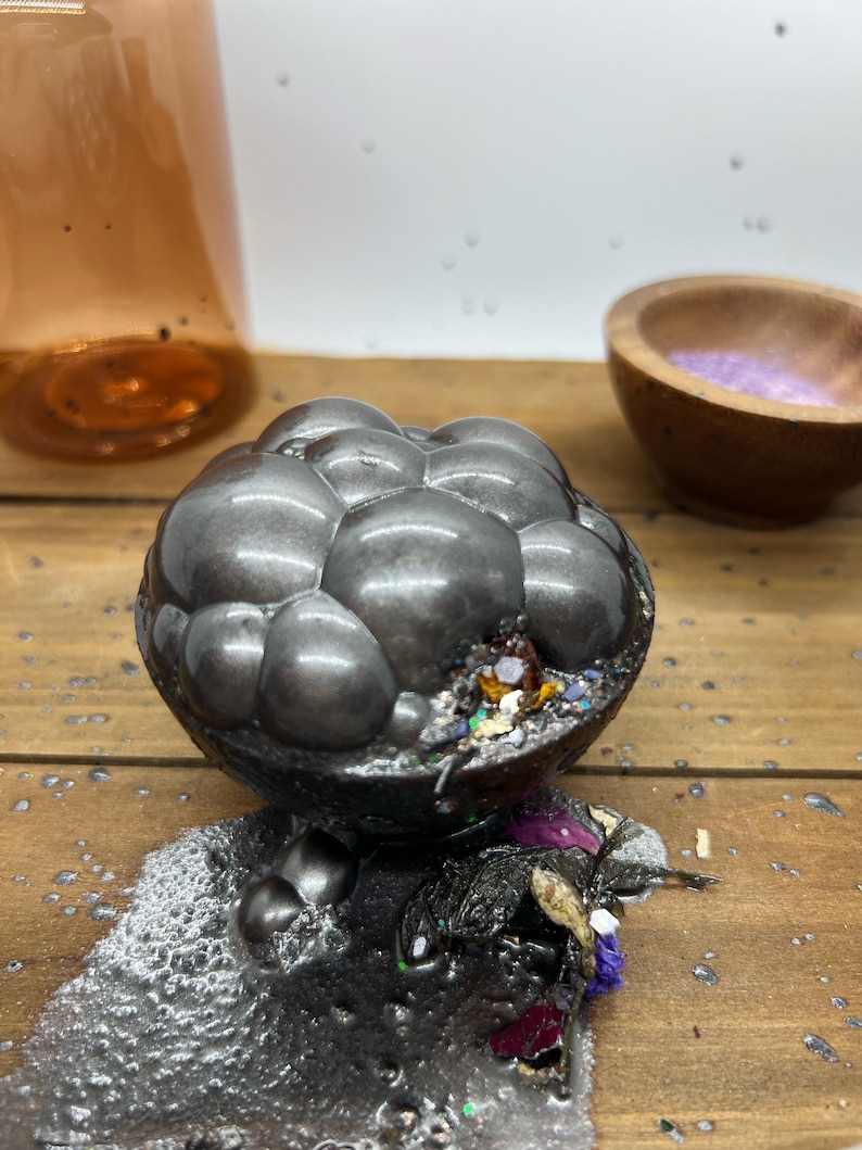 K&ouml;nnte beinhalten: Eine dunkelgraue, blasenartige Badekugel mit bunten Glitzer- und Trockenblumenakzenten. Die Badekugel liegt auf einer Holzoberfl&auml;che, mit einer Spur grauer R&uuml;ckst&auml;nde. Eine Glasflasche und eine Holzschale befinden sich im Hintergrund.