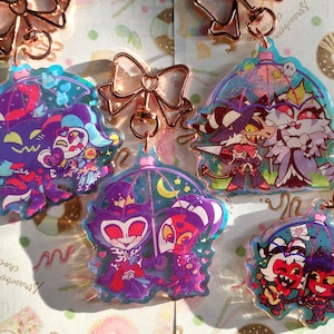 Puede incluir: Cuatro llaveros coloridos con personajes de dibujos animados bajo paraguas. Los personajes están en varias poses y tienen diferentes expresiones. Los llaveros están hechos de acrílico transparente y tienen un acabado holográfico.