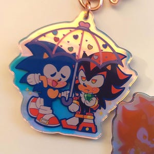 May include: A holographic charm featuring Sonic the Hedgehog and Shadow the Hedgehog sharing an umbrella with hearts on it. The charm is a light blue, dark blue, red, and black color.