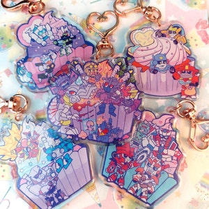 May include: A collection of colorful acrylic keychains featuring cartoon robot characters in cupcake designs. Each keychain has a rose gold clasp. The cupcakes have pink, blue, and purple frosting with sprinkles and stars.