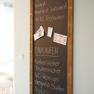 May include: A brown chalkboard with a wooden frame hangs on a light blue wall. The chalkboard is divided into two sections, one labeled "TERMINE" and the other labeled "EINKAUFEN". The "TERMINE" section lists appointments for a veterinarian, dermatologist, dentist, and a technician. The "EINKAUFEN" section lists items to buy, including a corkscrew, tissues, toilet cleaner, oregano, butter, and a pot cleaner.