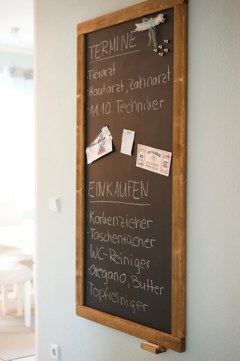 May include: A brown chalkboard with a wooden frame hangs on a light blue wall. The chalkboard is divided into two sections, one labeled "TERMINE" and the other labeled "EINKAUFEN". The "TERMINE" section lists appointments for a veterinarian, dermatologist, dentist, and a technician. The "EINKAUFEN" section lists items to buy, including a corkscrew, tissues, toilet cleaner, oregano, butter, and a pot cleaner.
