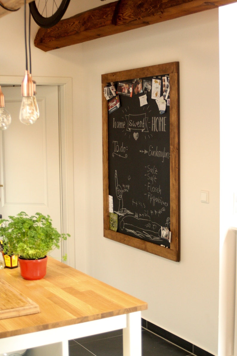May include: A wooden framed chalkboard with the words "home sweet home" written in white chalk. The chalkboard is decorated with photos, notes, and other items. The chalkboard is hanging on a white wall in a kitchen.