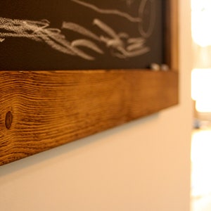May include: A close-up of a wooden frame with a dark brown stain. The frame is made of reclaimed wood with a knot in the center. The frame is mounted on a white wall.