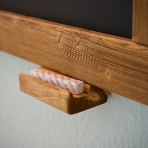 May include: A wooden chalkboard with a small wooden shelf for chalk. The shelf is attached to the bottom of the chalkboard and has a piece of chalk in it.