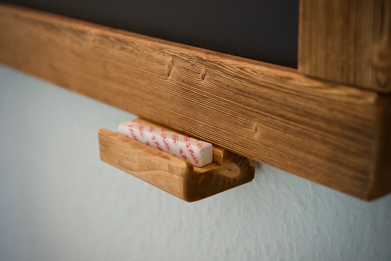 May include: A wooden chalkboard with a small wooden shelf for chalk. The shelf is attached to the bottom of the chalkboard and has a piece of chalk in it.
