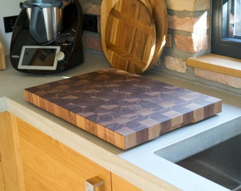 May include: A rectangular end-grain butcher block cutting board with a checkerboard pattern of light and dark wood. The board sits on a light-colored countertop near a stainless steel sink. Other wooden cutting boards are in the background.