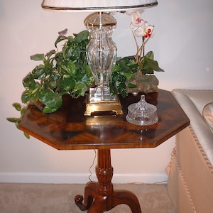 May include: A wooden octagonal table with a crystal lamp and a glass bowl on top. The table has three legs and a dark wood finish. The lamp has a white shade and a crystal base. The bowl is clear glass and has a decorative design.