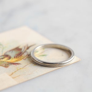 Puede incluir: Un anillo plateado con un diseño sencillo y elegante. Descansa sobre una postal vintage con una ilustración de pájaros. La banda del anillo tiene un sutil detalle de ranura, añadiendo un toque de sofisticación a la joya.
