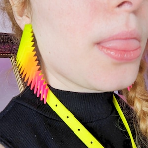 May include: A person wearing a black mock neck shirt with a yellow lanyard and a pair of bright pink and yellow earrings. The earrings are shaped like a jagged, pointed edge.