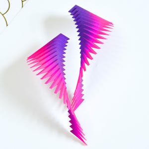 May include: A pair of pink and purple gradient earrings made of stacked paper triangles.