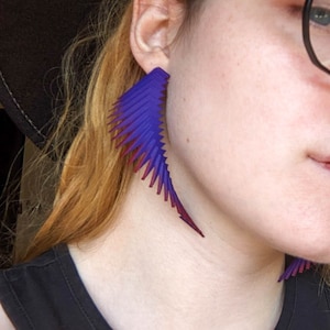May include: A pair of long, asymmetrical earrings with a gradient design. The earrings are made of a purple and red plastic material and have a sharp, pointed shape.