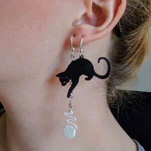 Pode incluir: Brincos de gato preto com um design de novelo de l&atilde; branco. Os brincos s&atilde;o feitos de um material escuro e apresentam uma silhueta de gato divertida. Os brincos s&atilde;o suspensos por ganchos prateados.
