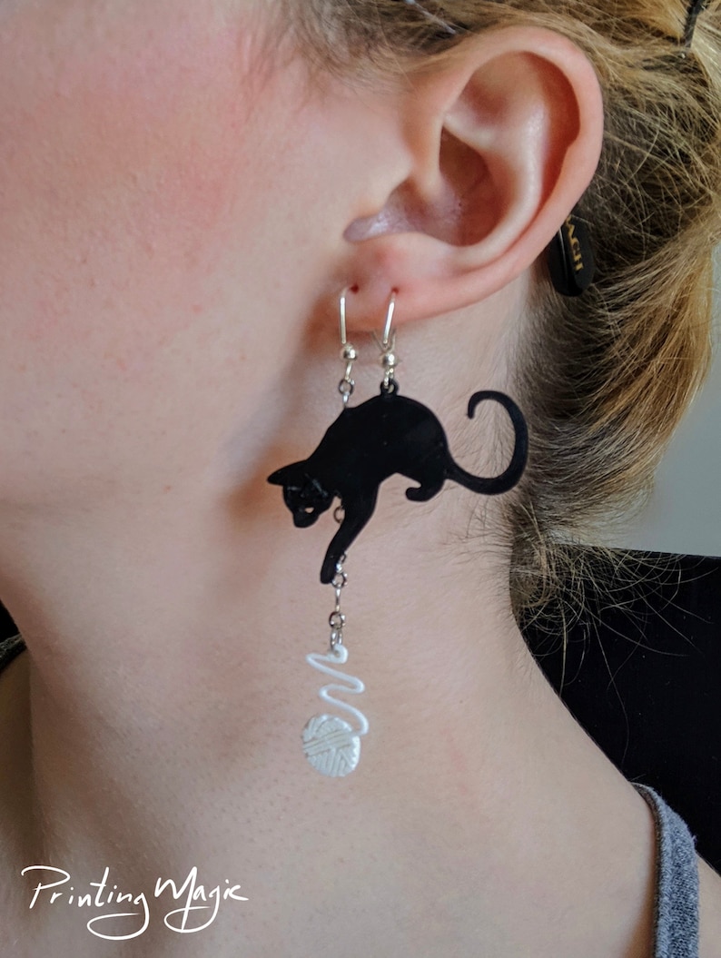 Pode incluir: Brincos de gato preto com um design de novelo de l&atilde; branco. Os brincos s&atilde;o feitos de um material escuro e apresentam uma silhueta de gato divertida. Os brincos s&atilde;o suspensos por ganchos prateados.