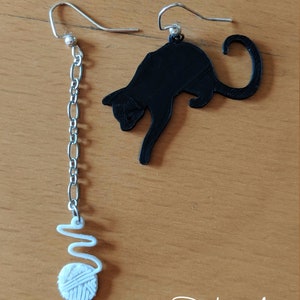 Pode incluir: Um par de brincos descombinados. Um &eacute; uma silhueta de gato preto, o outro apresenta uma corrente prateada com uma bola de l&atilde; branca e um redemoinho. Os brincos est&atilde;o sobre uma superf&iacute;cie castanha clara. O texto "Printing Magic" est&aacute; no canto inferior direito.