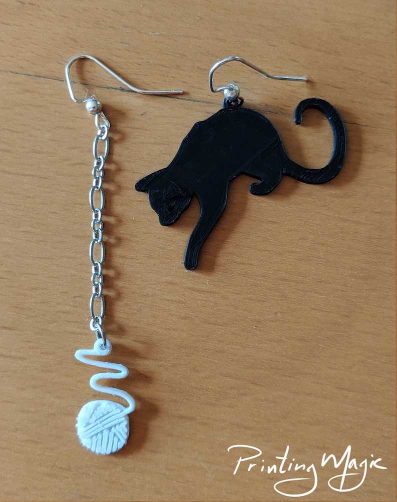Pode incluir: Um par de brincos descombinados. Um &eacute; uma silhueta de gato preto, o outro apresenta uma corrente prateada com uma bola de l&atilde; branca e um redemoinho. Os brincos est&atilde;o sobre uma superf&iacute;cie castanha clara. O texto "Printing Magic" est&aacute; no canto inferior direito.