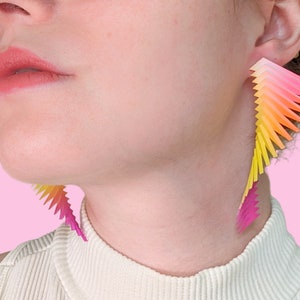 May include: A pair of long, colorful earrings with a gradient of yellow, orange, and pink. The earrings are made of thin, layered triangles that create a cascading effect.