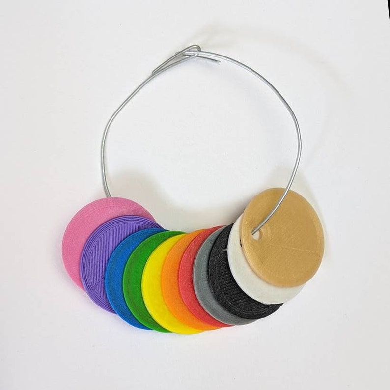 Pode incluir: Um anel de amostras de cores com uma variedade de amostras circulares de 2,5 cm de di&acirc;metro. As amostras est&atilde;o dispostas em um espectro de arco-&iacute;ris, incluindo rosa, roxo, azul, verde, amarelo, laranja, vermelho e outras cores. Um fio de prata prende as amostras.