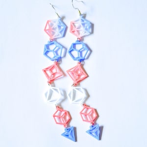 Puede incluir: Un par de pendientes largos y geométricos con un degradado de rosa, azul y blanco. Cada pendiente presenta una serie de colgantes en forma de cubo, calados y conectados entre sí.