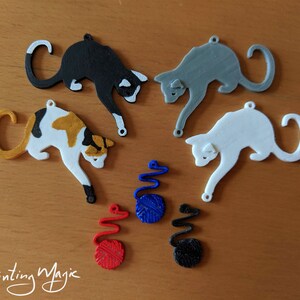 Pode incluir: Uma cole&ccedil;&atilde;o de enfeites em forma de gato em v&aacute;rias cores, incluindo preto, branco, cinza e tartaruga. Cada gato tem um pequeno orif&iacute;cio para pendurar. Tamb&eacute;m est&atilde;o inclu&iacute;dos enfeites de novelos de l&atilde; em azul, vermelho e preto. O texto "Printing Magic" est&aacute; na parte inferior.