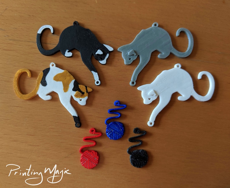 Pode incluir: Uma cole&ccedil;&atilde;o de enfeites em forma de gato em v&aacute;rias cores, incluindo preto, branco, cinza e tartaruga. Cada gato tem um pequeno orif&iacute;cio para pendurar. Tamb&eacute;m est&atilde;o inclu&iacute;dos enfeites de novelos de l&atilde; em azul, vermelho e preto. O texto "Printing Magic" est&aacute; na parte inferior.