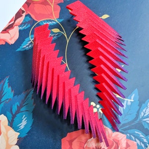 May include: Two red and purple ombre geometric shapes made of layered paper. The shapes are arranged in a curved pattern.