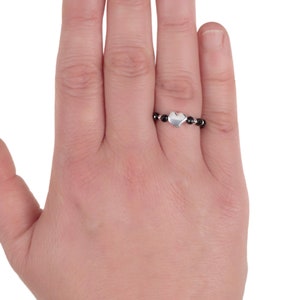 May include: A silver heart charm ring with black beads. The ring is on a person's finger.