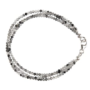 Könnte beinhalten: Eine zarte Halskette mit drei Strängen aus kleinen, facettierten Perlen in den Farbtönen Klar, Grau und Schwarz. Die Halskette wird mit einem silberfarbenen Verschluss geschlossen. Die Perlen sind gleichmäßig angeordnet und wirken raffiniert und elegant.