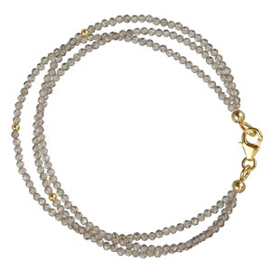 Puede incluir: Un brazalete de triple cadena con cuentas de cristal gris y detalles dorados. El brazalete tiene un cierre dorado.