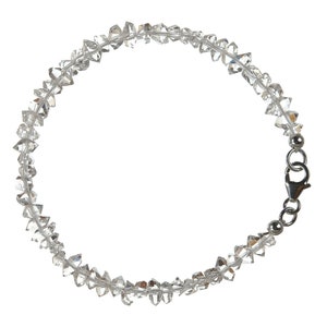 Könnte beinhalten: Ein Armband aus klarem Quarz-Kristall. Das Armband besteht aus unregelmäßig geformten, klaren Quarz-Kristallen, die aufgefädelt sind. Ein silberfarbener Verschluss sichert das Armband. Die Kristalle sind facettiert und fangen das Licht ein. Das Armband ist für das Handgelenk konzipiert.