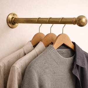 Peut inclure: Un portant à vêtements en laiton avec trois cintres en bois portant une chemise de couleur claire, un pull gris et une chemise bleu foncé. Le portant est fixé sur un mur de couleur claire.