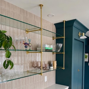 May include: A wall-mounted shelving unit featuring three horizontal glass shelves supported by vertical brass poles. The shelves hold various clear and tinted glassware, a green-spined book, and a small potted plant, set against a beige tiled wall.