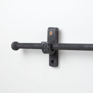 May include: A black metal towel bar mounted on a white wall. The bar has a cylindrical design with a ball-shaped end. The mounting hardware is also black and includes a bracket and screws. The overall style is rustic and industrial.
