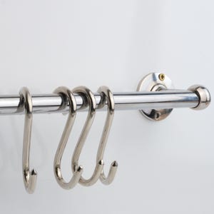 Könnte beinhalten: Ein Küchenutensilienhalter aus Edelstahl mit vier S-förmigen Haken. Der Halter ist an einer weißen Wand montiert. Die Haken sind zum Aufhängen von Küchenutensilien, Töpfen und Pfannen gedacht. Der Halter ist aus glänzendem, poliertem Metall gefertigt.