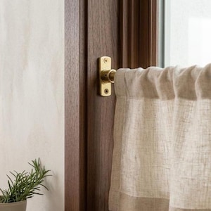 Puede incluir: Primer plano de una ventana de cocina con un tirador de latón y una cortina de lino beige. El marco de la ventana es de color marrón oscuro y la pared es de color beige claro. Una pequeña planta de romero está en primer plano.