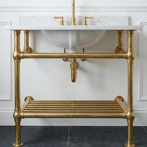 以下が含まれることがあります： 白い大理石のカウンタートップと白い磁器の洗面台を備えた、金色の金属製のバスルームバニティ。 バニティには棚と配管の詳細があります。 蛇口とハンドルも金色です。