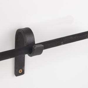 May include: A black, wrought iron towel bar mounted on a white wall. The bar has a rounded end and is held in place by a vertical bracket with a curved top and a small, square hook. A single screw secures the bracket.