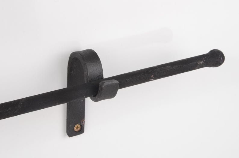 May include: A black, wrought iron towel bar mounted on a white wall. The bar has a rounded end and is held in place by a vertical bracket with a curved top and a small, square hook. A single screw secures the bracket.