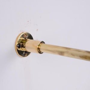 May include: A brass-colored curtain rod, mounted on a white wall. The rod is cylindrical and has a slightly aged appearance. The mounting hardware includes a circular plate with screw holes.
