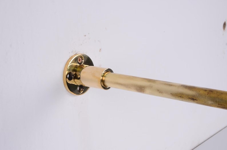 May include: A brass-colored curtain rod, mounted on a white wall. The rod is cylindrical and has a slightly aged appearance. The mounting hardware includes a circular plate with screw holes.