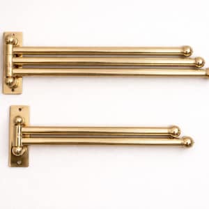 Peut inclure: Deux porte-serviettes en laiton montés sur un mur blanc. Chaque porte-serviettes a une base rectangulaire avec des trous de montage et plusieurs barres horizontales qui pivotent. Les barres se terminent par des embouts arrondis en forme de boule. Le laiton a une finition polie.