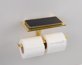 Könnte beinhalten: Ein goldfarbener, an der Wand befestigter Toilettenpapierhalter mit einer Ablage für ein Telefon. Der Halter hat zwei Rollen weißes Toilettenpapier. Das Telefon ist schwarz. Der Halter ist aus Metall.